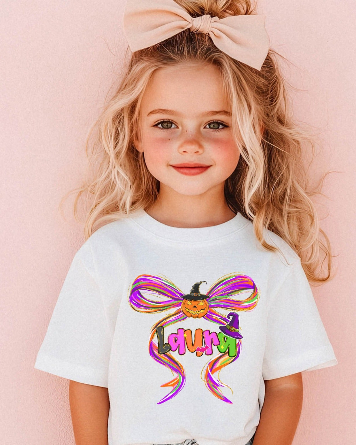 Little girl wearing a white t-shirt with a colorful Halloween bow design featuring a pumpkin, witch hat, and vibrant ribbon accents.