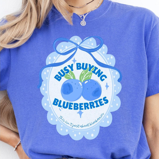 Periwinkle blue graphic tee with scalloped frame design, blueberries, and the words "Busy Buying Blueberries" accented with a bow.