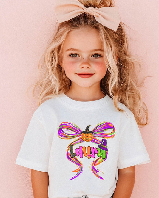 Little girl wearing a white t-shirt with a colorful Halloween bow design featuring a pumpkin, witch hat, and vibrant ribbon accents.